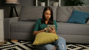 Attractive young woman taping on the tablet while sitting on the floor in the cosy nice living room. Home