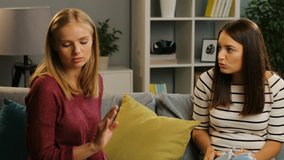 Upset blonde young woman telling something sad to her brunette female friend in the nice living room. Inside