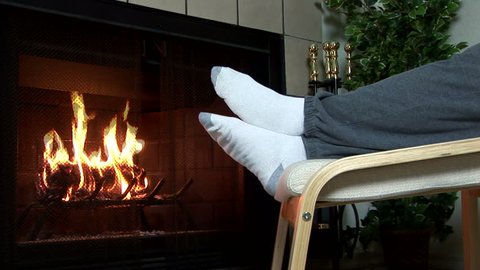 A man sitting next to his living room fireplace warming his feet by the cozy fire. 