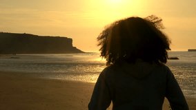 4K video clip of sad thoughtful mixed race African American girl teenager female young woman with curly hair standing on a beach looking at sunset or sunrise