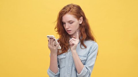 Happy surprised ginger woman in denim shirt using her smartphone over yellow background
