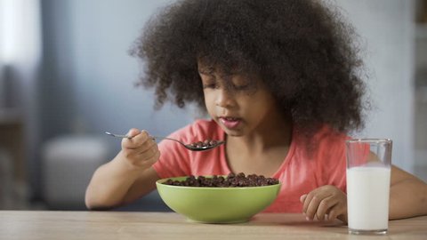 Pretty African American girl eating cereal with appetite and drinking milk