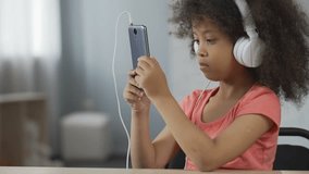 Serious African-American kid wearing headset and watching video on cellphone