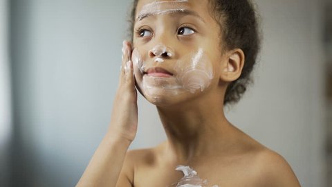 Funny Afro-American kid putting moisture cream on face, spa procedure, beauty