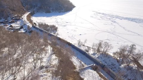 From above top passengers train Trans Siberian railway old village. Frozen lake Baikal coast. Winter beautiful Holiday  Russia. Sunny day snow field high rocks. Fast speed aerial drone 4k footage.