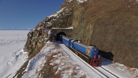 Passengers train Trans Siberian railway from tunnel. Frozen lake Baikal coast. Winter beautiful Holiday Russia. Sunny day snow field high rocks. Fast speed aerial drone 4k footage.