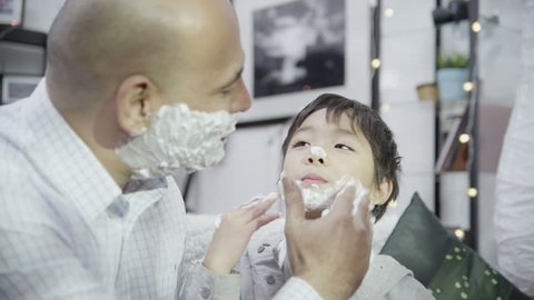 Loving father teaches his cute young son how to apply shaving foam to his face. In slow motion.