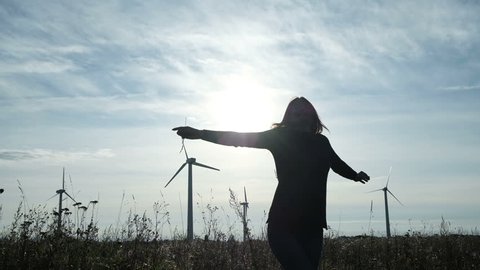 One free pretty lady enjoying turn with opening arms on the meadow slow motion. The weather is natural sunny, cloudy and windy, skyline at horizon. Background eco electricity windfarm with propellers