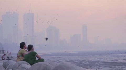 Two middle aged women sitting on tetra-pods and chatting next to the seashore. while group of crows flying and city skyline covered in smog visible in background in the morning, Mumbai, India (2017)