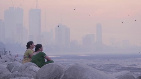 Two middle aged women sitting on tetra-pods and chatting next to the seashore. While city skyline visible in background in the morning, Mumbai, India (2017)