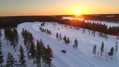 AERIAL: Flying above the car driving through snowy forest at golden winter sunrise. People on winter road trip traveling across snow covered Lapland wilderness at sunset. Car driving on empty icy road