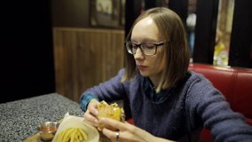 Young girl is eating untasty burger and French fries in the cafe. A woman in glasses is disappointed with the taste of stale cutlets in a hamburger and feel bad. Fast food. Closeup shot.