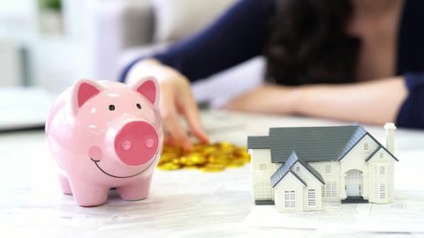 lovely young woman sitting in living room  at home and holding gold coin saving money into pink piggy bank for buying new house