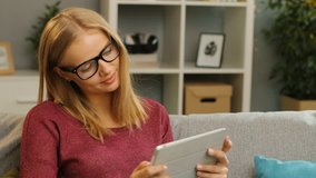 Pretty young blonde woman in glasses scrolling and looking carefully on the tablet screen on the couch in the cosy living room. Portrait shot. Inside