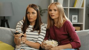 Two frightened pretty young women eating popcorn on the couch, watching horror. Blonde and brunette in the cosy living room. Indoor