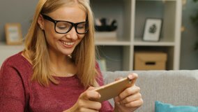 Smiled young woman in glasses exciting by playing game on smart phone while sitting on the sofa in the modern living room. Portrait shot. home