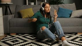 Pretty young woman in the white headphones listening to the music on the smart phone, singing and dancing in the nice living room on the floor. Indoor