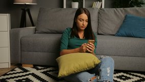 Attractive young woman taping on the smart phone and sitting on the floor in the cosy living room. Indoor