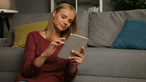 Beautiful blonde young woman scrolling the smart phone and smiling. Girl sitting on the floor in the cosy nice room. Indoor