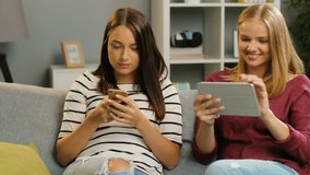 Two pretty young women sitting on the couch with tablet device and smart phone, laughing and showing something to each other. Blonde and brunette in the modern living room. Home
