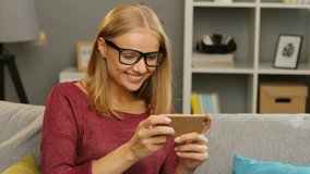 Beautiful young blonde woman in glasses playing game on smart phone and winning while sitting on the sofa in the nice living room. Home