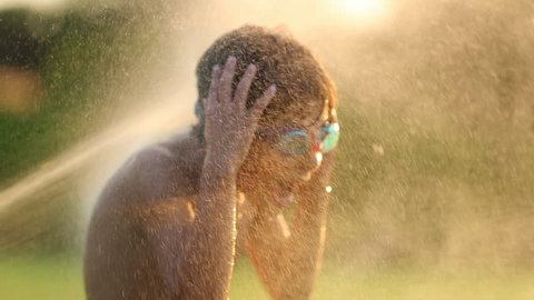 Child having fun with water sprinkles in the lawn during sunset golden hour time. Child yelling dramatically while water splashes him at 120fps. Cinematic dramatic super slow-motion scene