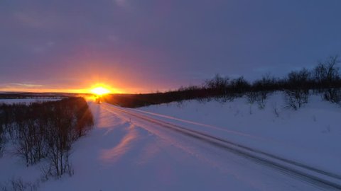 AERIAL: Trucking at winter sunrise. Semi truck transporting goods at sunrise. Speeding heavyweight lorry emerging from golden sunshine during sunset on a snow covered highway surrounded by bare trees.