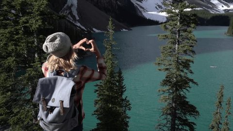 Slow motion shot of young woman standing looking at stunning mountain lake views makes a heart shape frame using her fingers.
Woman loving nature at Moraine lake, Banff, Alberta, Canada