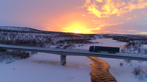 AERIAL: Flying above semi truck driving along scenic countryside highway at sunset. Transporter lorry crossing a bridge above frozen icy river in picturesque snowy landscape at gorgeous golden sunrise