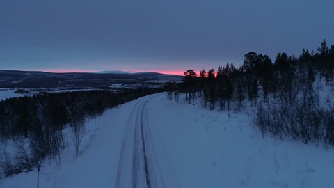 AERIAL: Flying above empty mountain highway running through snowy winter wilderness in Lapland, Finland at sunrise. Icy rutted road on a hill in wintry countryside at beautiful pink and orange sunset