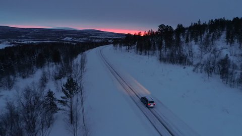 AERIAL: Flying above car driving along empty icy mountain highway through snowy spruce forest at golden winter sunset. People on road trip traveling across snow covered Lapland wilderness at sunrise