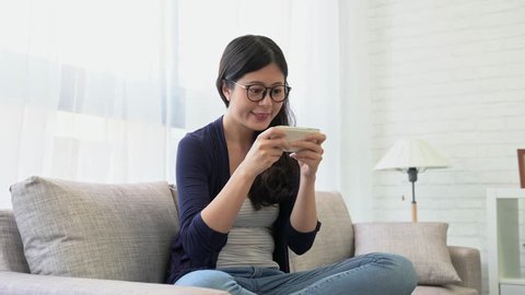 Dolly shot of beautiful woman playing smartphone app game and celebrate winning. happy girl text messaging application while sitting on sofa at home. 