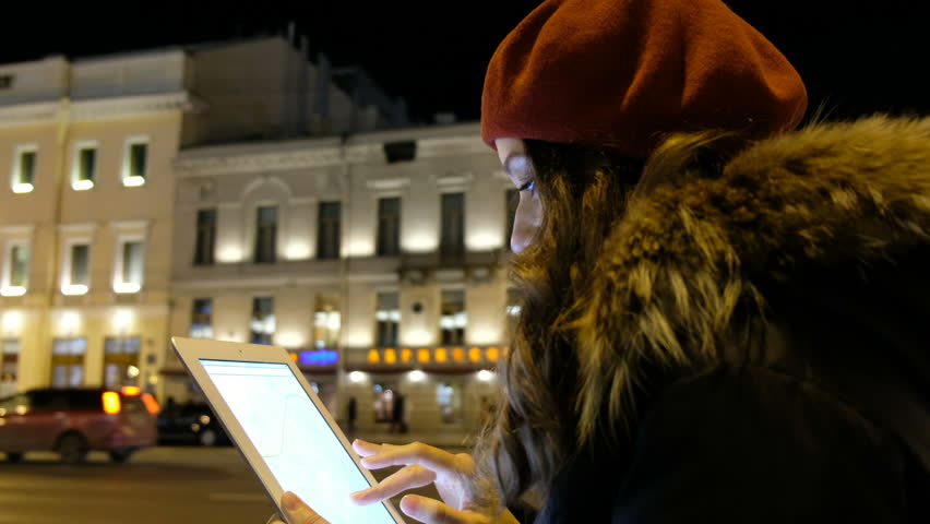 Attractive girl looks out the tablet and waiting for the bus in the evening in the city.