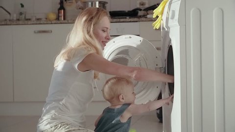 Young mother with baby sitting on floor front washing machine and putting dirty laundry into. Young mother using washing machine for wash