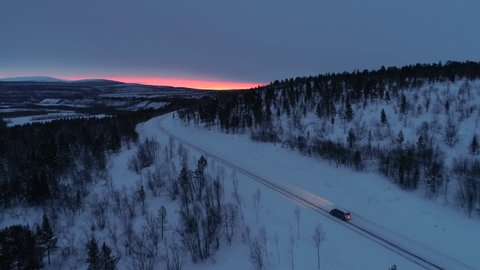 AERIAL: Flying above car driving in dark along empty mountain highway through snowy spruce forest at golden winter sunset. People on road trip traveling across Lapland wilderness in Finland at sunrise