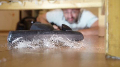 Middle-aged man cleaning a floor under a bed lying on the floor using vacuum cleaner