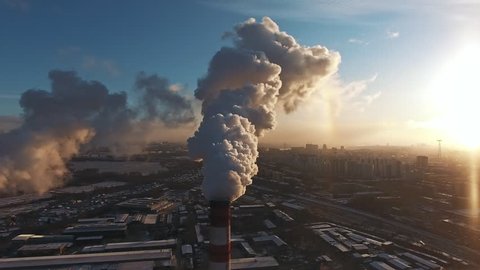Three industrial white and red pipes smoke against contrasting sun. Thick, dense smoke comes from the pipe, removed from quadrocopter. Fly over an industrial area with working pipes, smoke is pouring.
