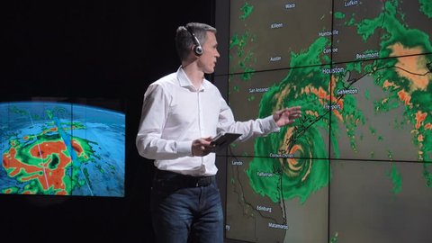 A news weather reporter wearing a headset presenting a live hurricane forecast displayed on two large screens. Elements of this image furnished by NASA.