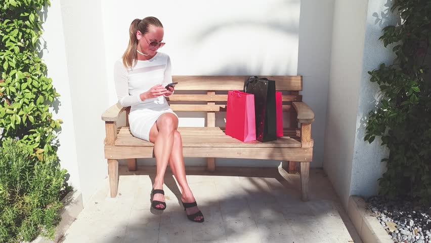 Slow motion. Beautiful woman in sun glasses are holding shopping bags, using a smart phone and smiling