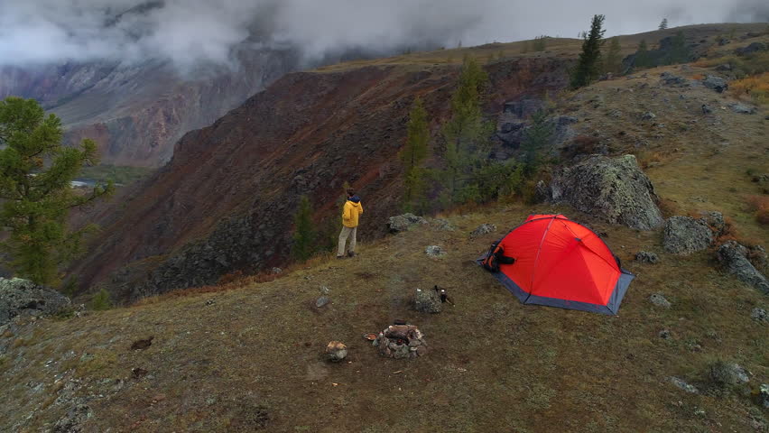 aerial footage of a man standing near a tent in front of mountain valley. flying around. cloudy sky.