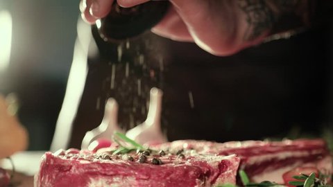 Male tattooed Chef’s Hand Seasoning Fresh steak in a Bright Light. Cuisine, Bon Apetite, Fresh Meat, Pepper Salt, Garlic and Rosemary. Close Up View, Slow Motion.