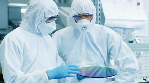 Two Scientists/ Technicians in Sterile Suits Check Semiconductor Silicon Wafer that Reflects Many Different Colors, it will be Made into Computer Chips.They Work in a Semiconductor Manufacturing Plant