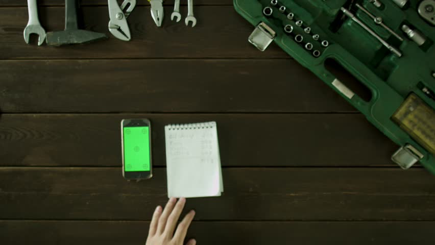 A man sits at a table near toolbox and writes something in notebook from phone.