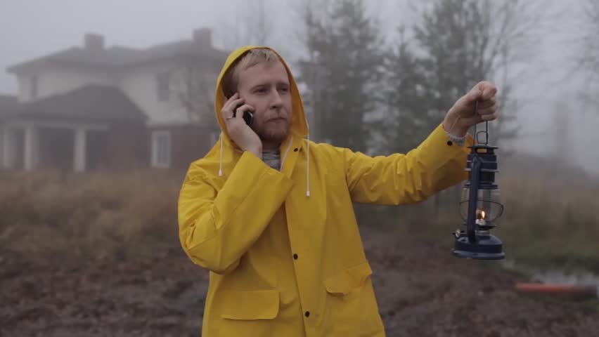 man in a yellow raincoat talking on the phone with a lantern by the lake in a fog