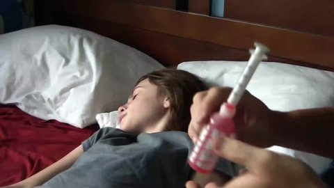 Clip of sick girl in bed given oral syringe of medicine, administered by parent.