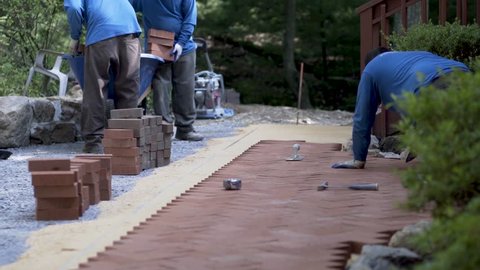 As men move piles of red bricks into the paving area, another man uses a trowel to get space between the brick pavers.