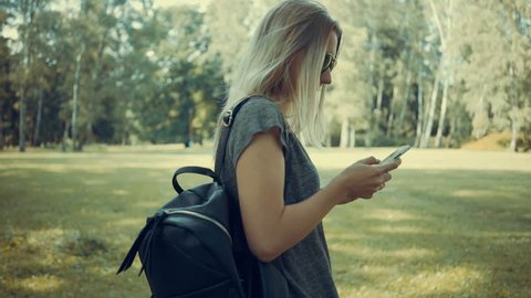 Woman walking in park with her phone at summer day under sun light. Slow motion