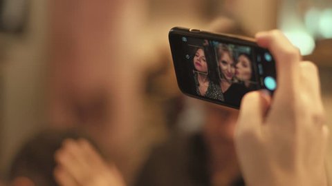 Soft focus hand held shot of three cute pretty girls, hipster millennials from young generation make selfie photo on modern frontal camera of smartphone during party, to share on social media