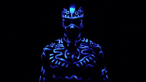A black man in fluorescent patterns dances in ultraviolet light, slow motion