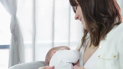 Young mother feeding breast her baby at home in white room
